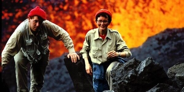 A man and woman smile in front of an erupting volcano