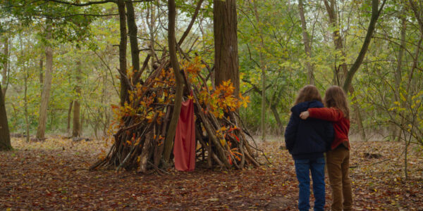 Two little girls seen from behind in the wood looking at a fort made of sticks