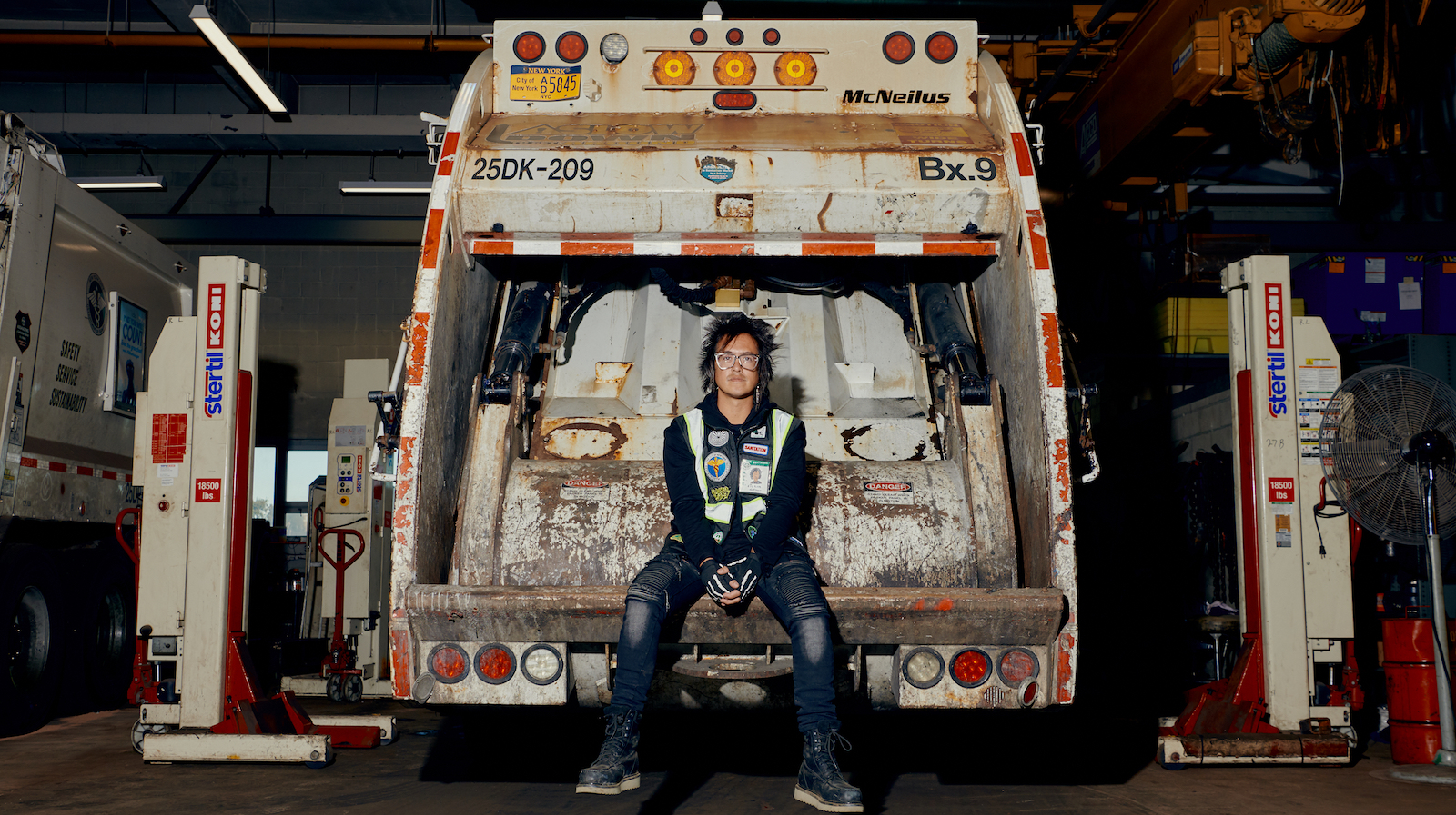 A man in glasses sits in the back of a garbage truck in a garage looking at camera