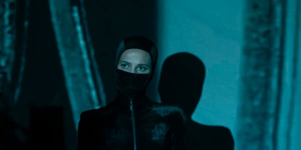 A woman in a zip-up black leather suit and the bottom of her face obscured by a mask looks past the camera; her shadow on the wall behind her