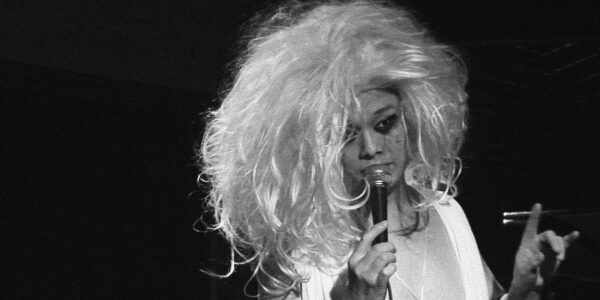 a drag performer with a white shock wig talks into a microphone with one finger pointed up