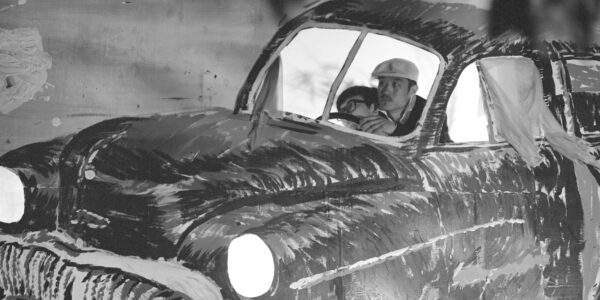 two men sit in the driver's seat of a cardboard illustrated car