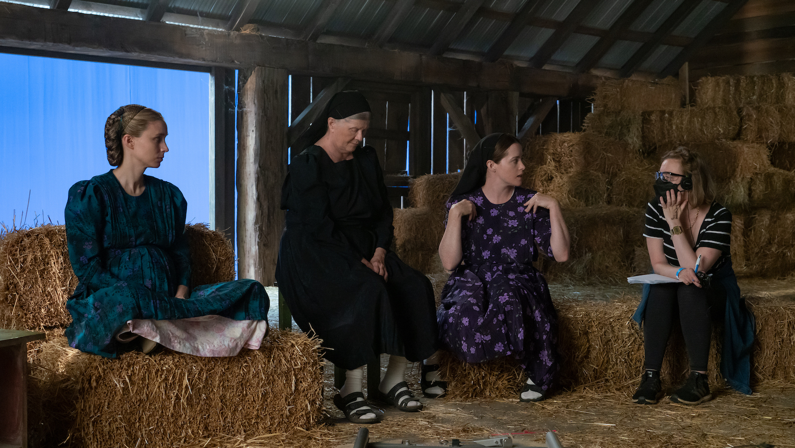 Four women sitting in a hayloft in a barn talk with each other, including the director on the right, listening with a pen and paper and wearing a mask and headphones