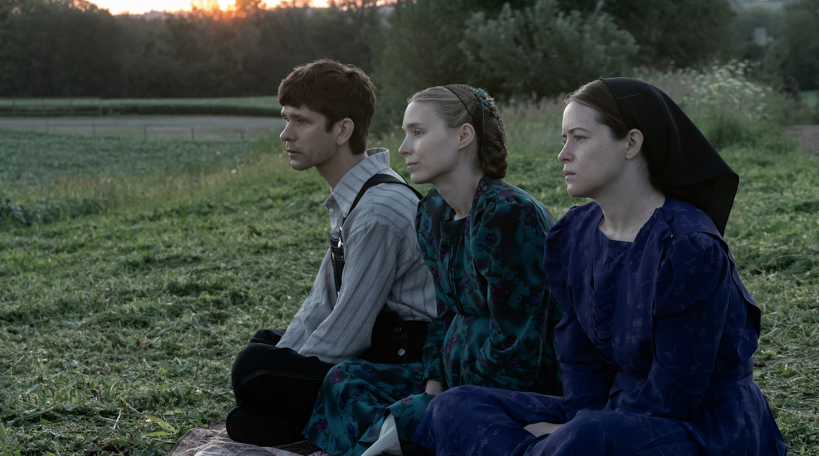 A man and two women sit in a field and look to the right off camera before a rising morning sun