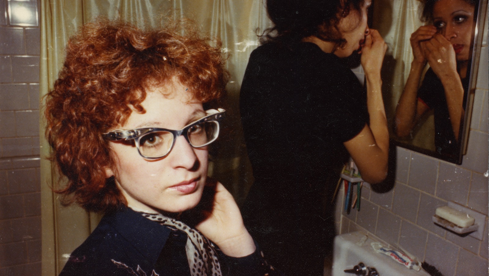 A woman in glasses and frizzy red hair looks in the camera away from a mirror
