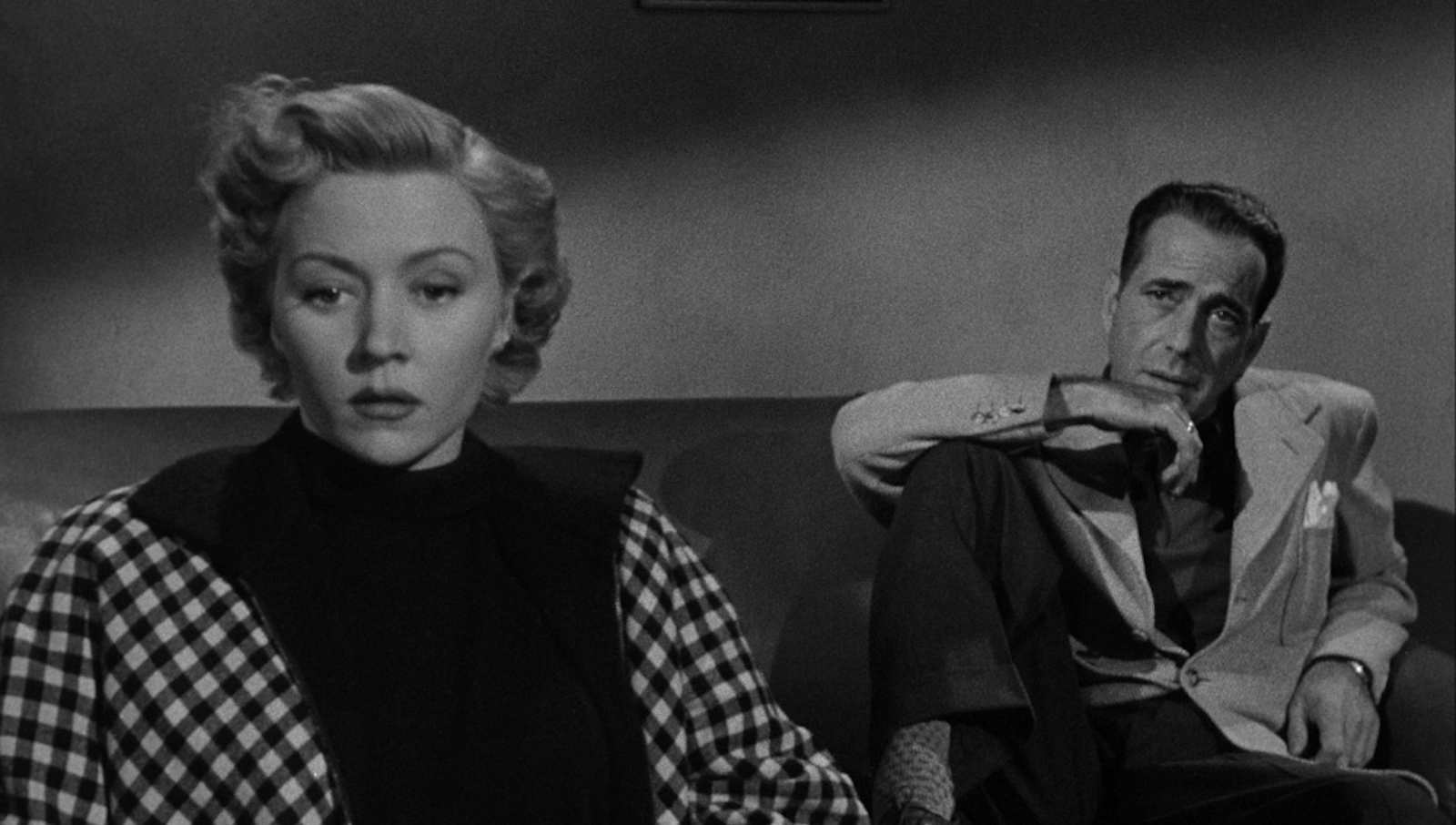A woman in the foreground looks off screen concerned, while a man sits behind her on the same couch looking at her