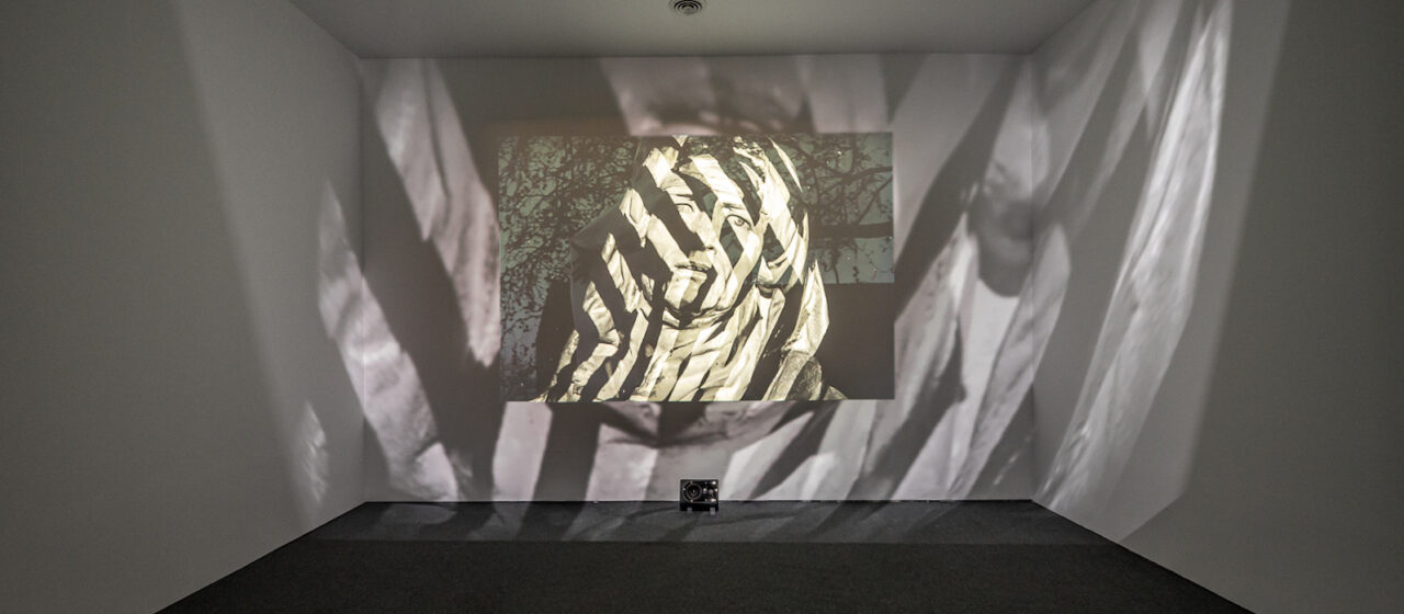 A gallery with a screen showing a statue's face, over which is projected black and white stripes that also emerge out of the screen onto the walls of the gallery