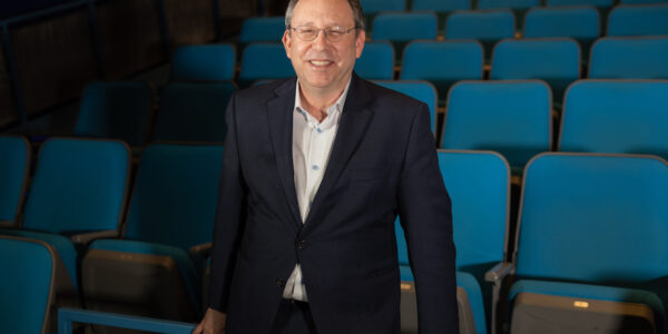 a man in a navy blazer stands in a theater with blue seats
