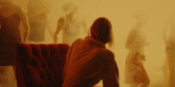 A woman seen from the back sits in a smokey yellow room and watches other women dance in the fog