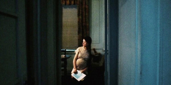 a pregnant woman sits alone, holding a piece of paper in a vacant room, shot from a distance