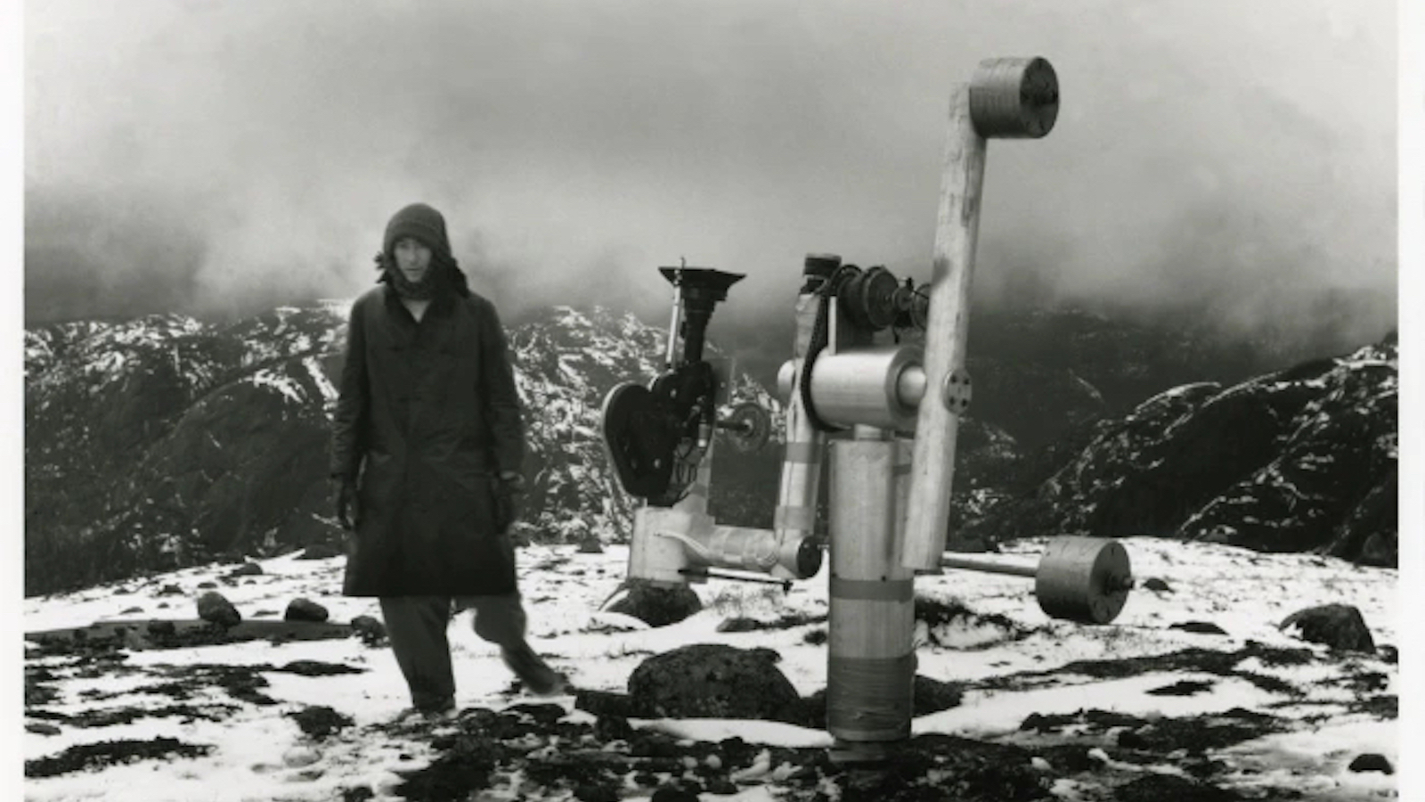 A man stands on a mountaintop next to an apparatus with a telescope
