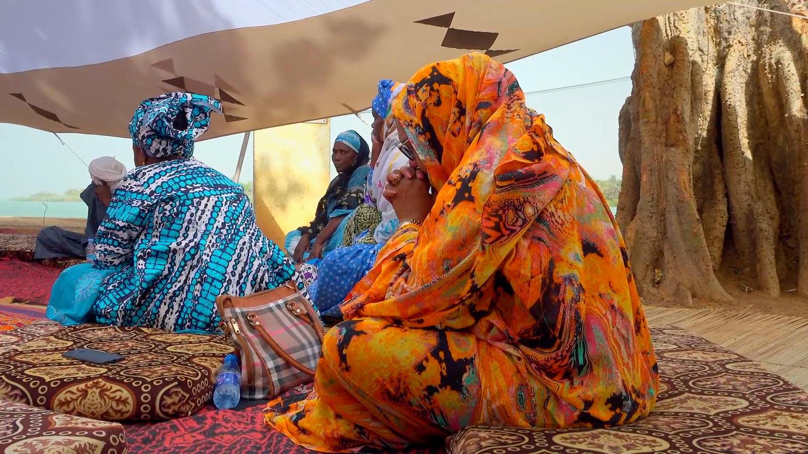 Women in brightly colored orange and blue saris sit cross legged on a mat under a tent outside