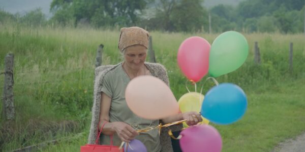 An elderly woman in a rural, green area stands by the roadside holding a cluster of colorful balloons