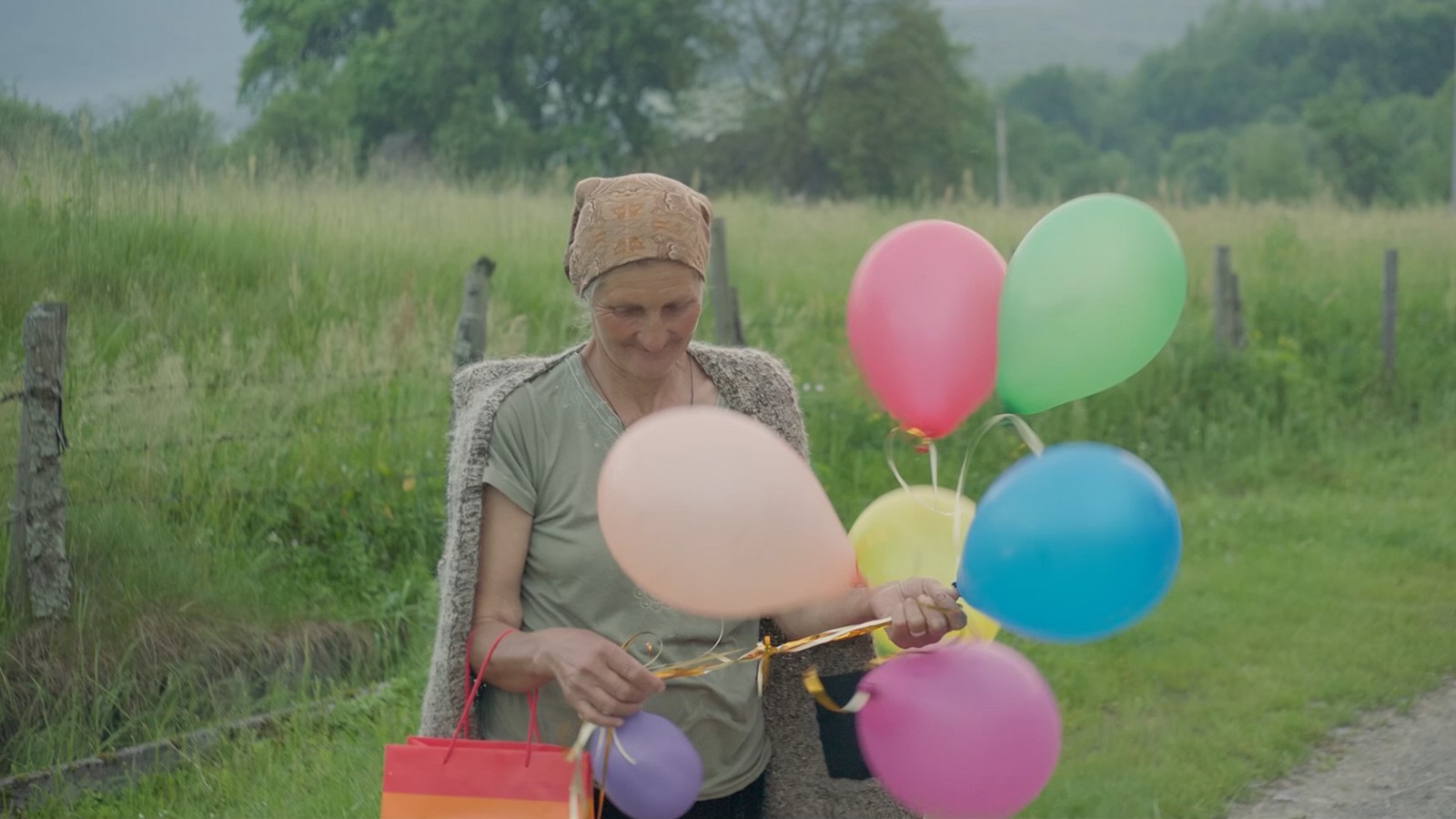 An elderly woman in a rural, green area stands by the roadside holding a cluster of colorful balloons