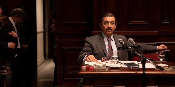 A mustachioed man sits in a dark room with wood paneling in front of a microphone, lit from above