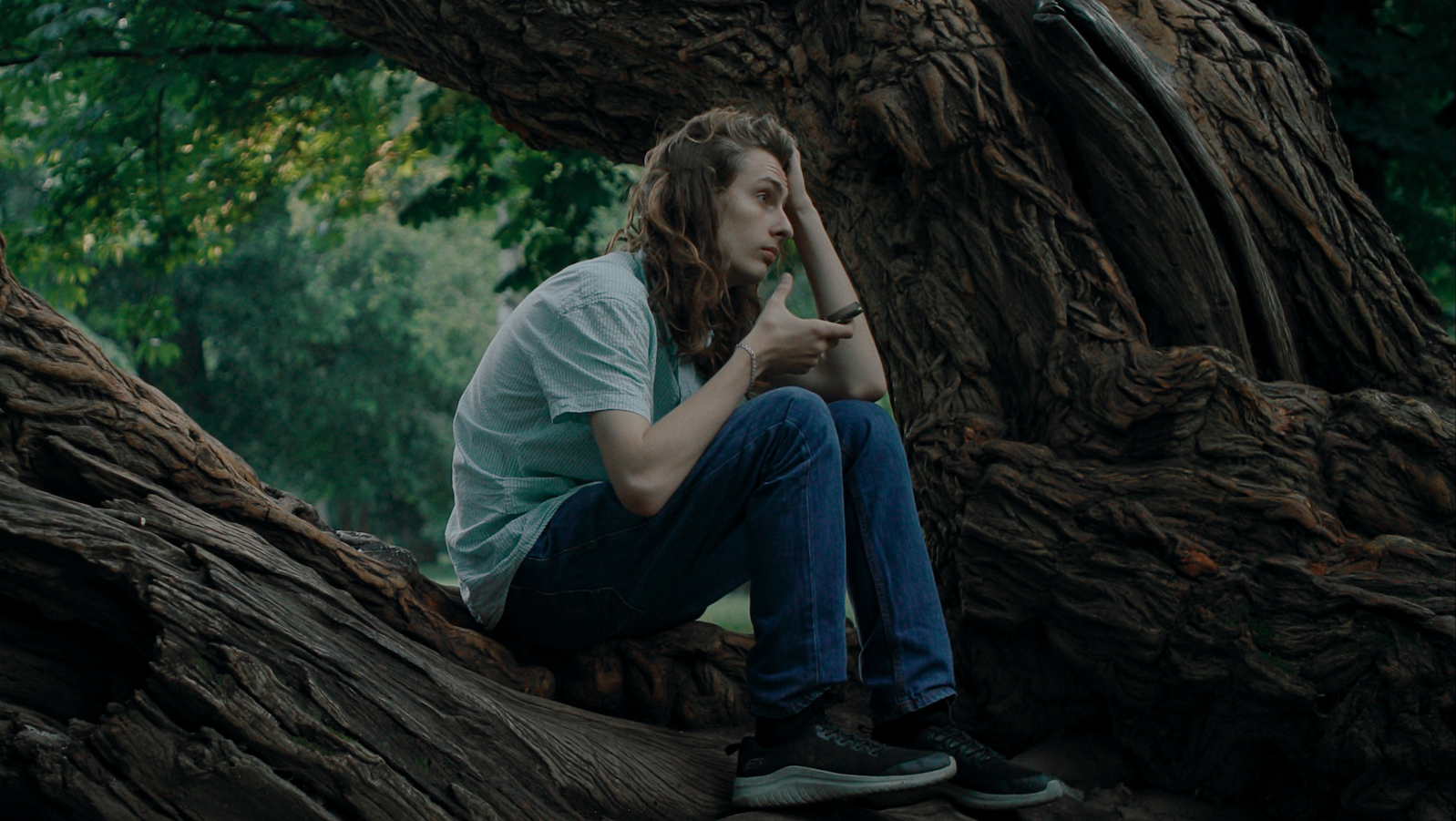 A young person with long hair sits on a tree in a forest holding a phone and looking into the distance