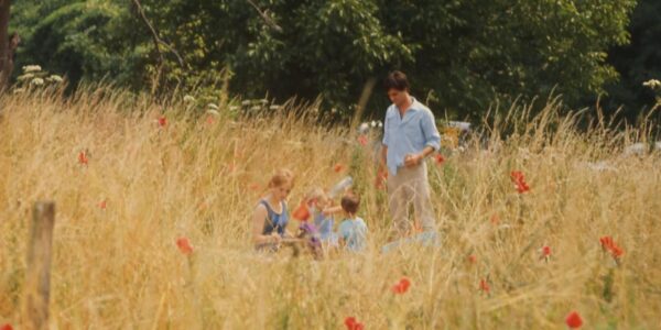 A man stands in a yellow field next to a woman and a young child, surrounded by red summer flowers