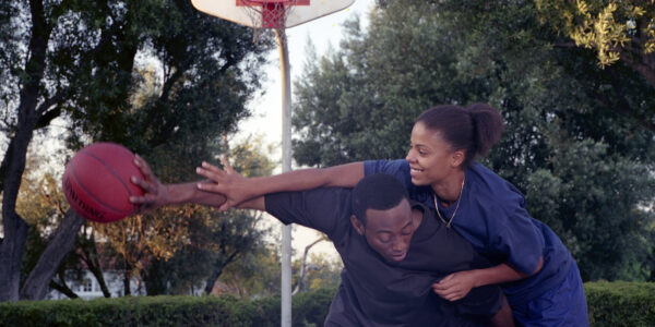 A young man and woman play basketball at an outdoor court, she's trying to block him while smiling