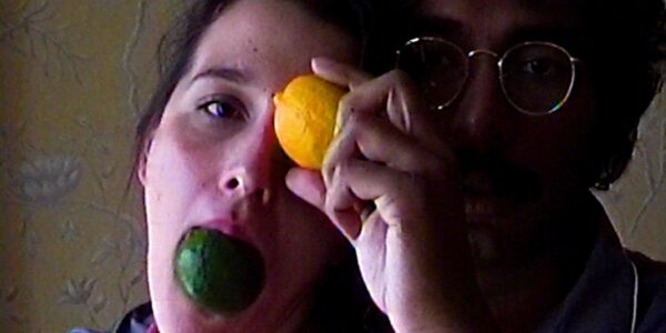 A woman holds a lime in her mouth and a lemon against her eye, with a bespectacled man behind her