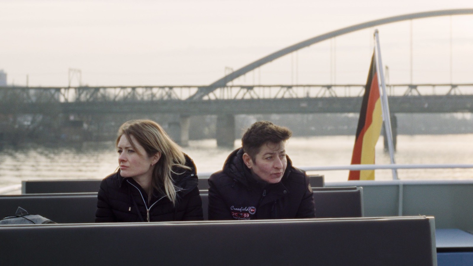 Two women sit on a ferry, huddled in the cold, a bridge in the background