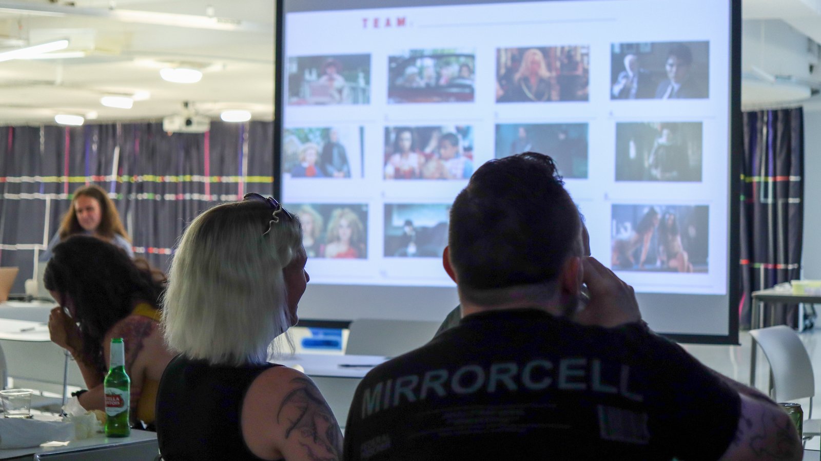 From the back, a man and a woman playing trivia sit in front of a screen with small images from films on it.