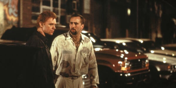 Two men with intense expressions stand in a parking lot; the man with a goatee looks intently at the red-headed man, looking away.