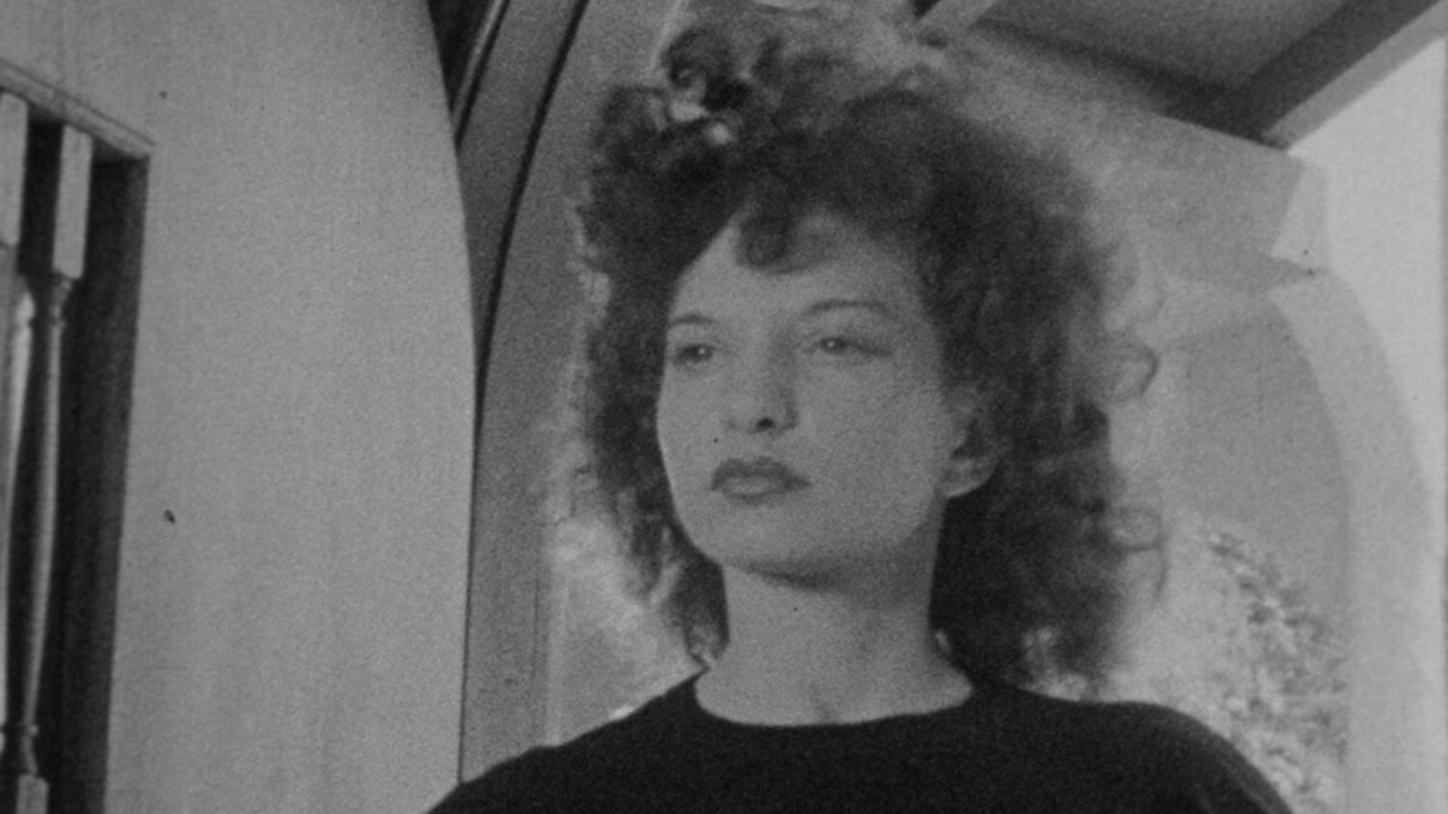 Standing inside a house, a woman with curly brown hair looks off into the distance past the camera