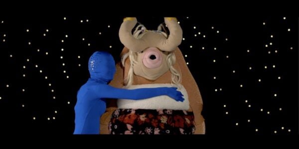 A figure wrapped entirely in blue head, including their head, wraps its arms around a large puppet-like creature with horns and an open mouth facing camera, with a sea of stars behind them