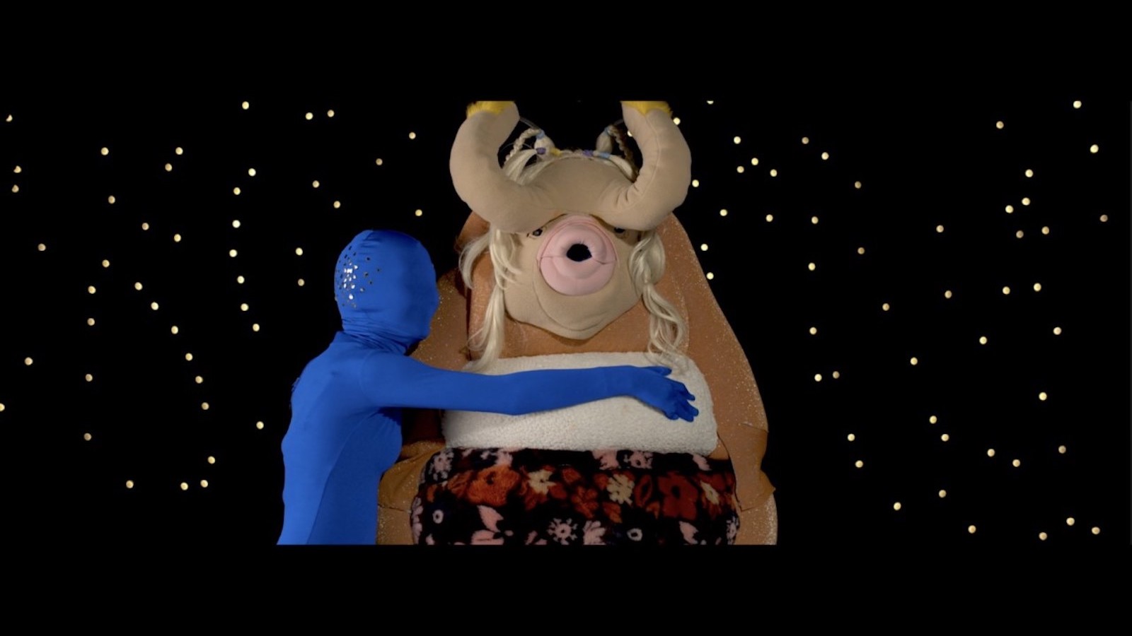 A figure wrapped entirely in blue head, including their head, wraps its arms around a large puppet-like creature with horns and an open mouth facing camera, with a sea of stars behind them
