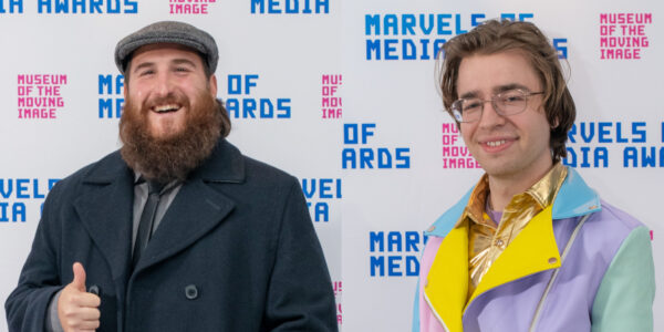 A bearded person in a coat smiles and gives the thumbs up and another wearing a multicolored jacket and glasses smiles, both with a Museum and Marvels of Media step-and-repeat in the background