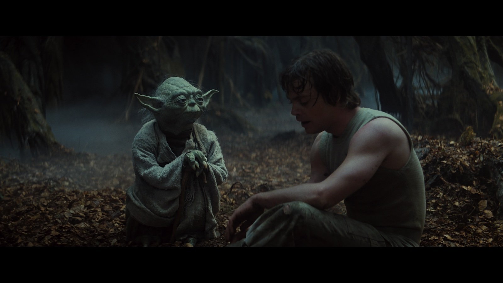 A small green creature named Yoda leans on a cane as he looks at a man in a tank top in a wooded area