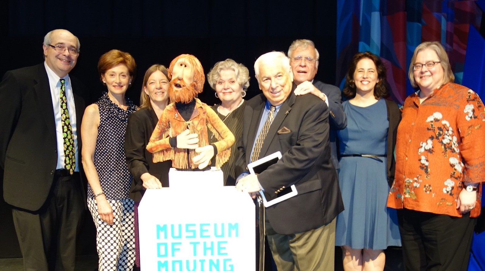 A bearded muppet likeness of Jim Henson stands at a Museum stage podium, surrounded by eight people looking at camera.