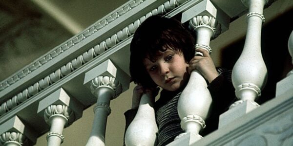 A little mop haired boy pokes his head through staircase railings and looks down