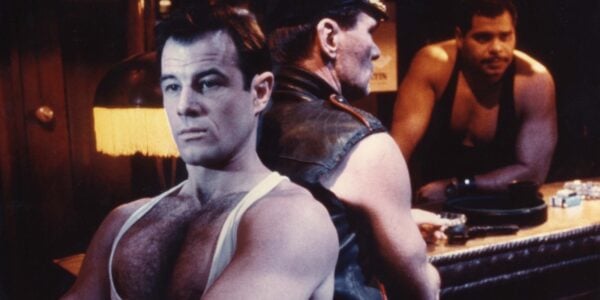 A man in a low cut tank top sits with his arms crossed, his back to another man wearing a leather cap and vest in a bar, with a bartender watching from behind them