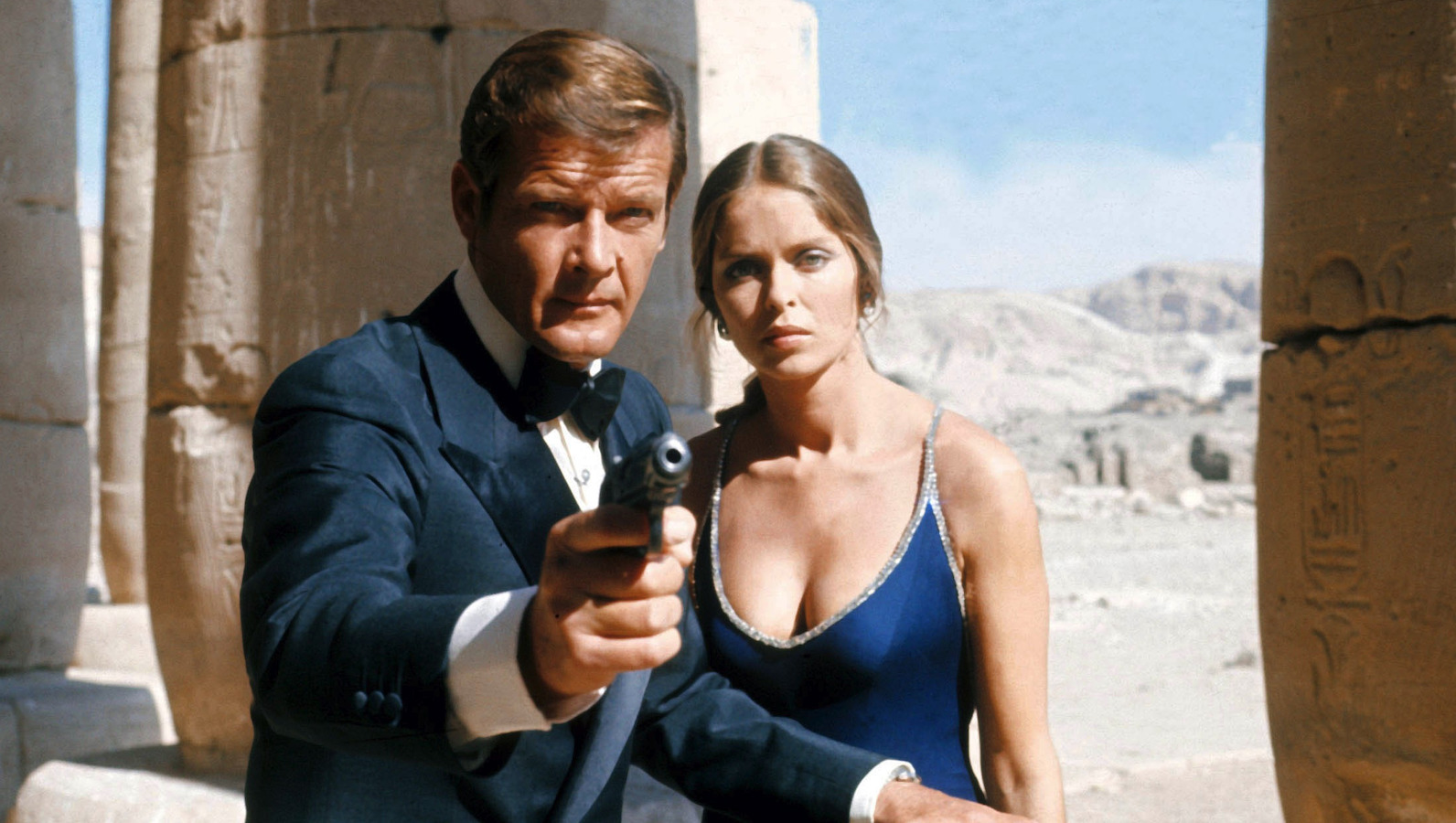 A man in a tux holding a gun with a blonde in a low cut dress behind him