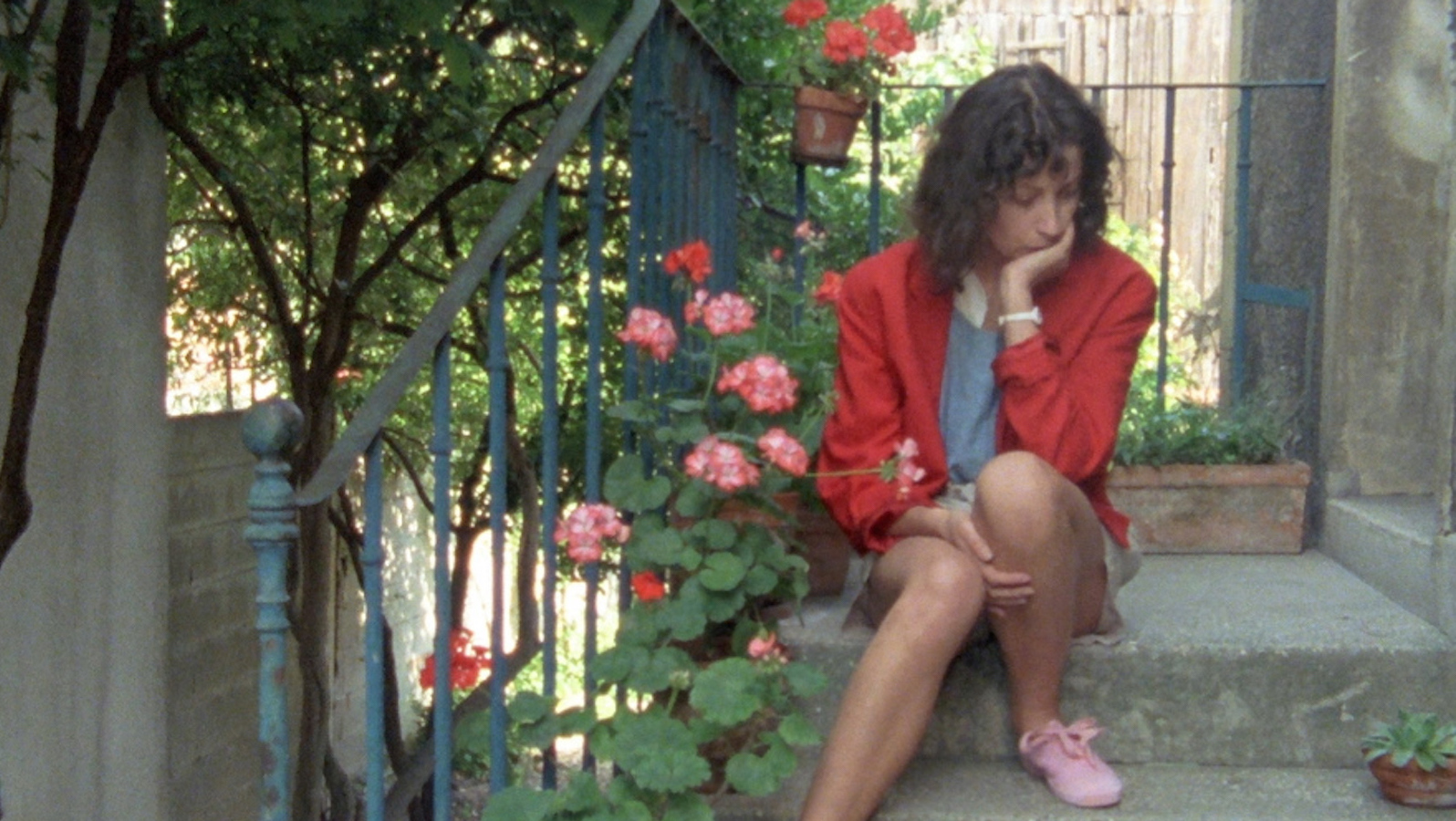 A woman in red jacket sits on outdoor steps next to pink flowers while she looks down, her chin resting in her hand