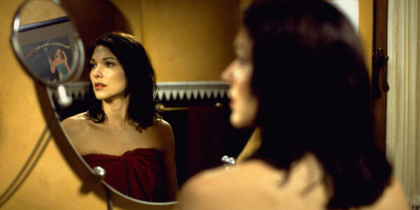 A dark haired woman in a red towel looks in a bathroom mirror at an image of a woman on a movie poster