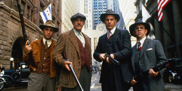 Four men in hats and 1920s garb hold shotguns and look at camera