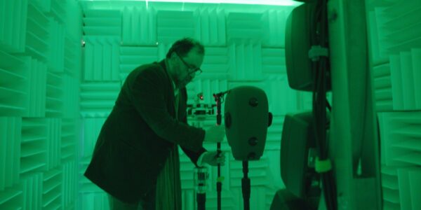 A man in glasses holds a microphone stand in a sound-proofed room all drenched in green light