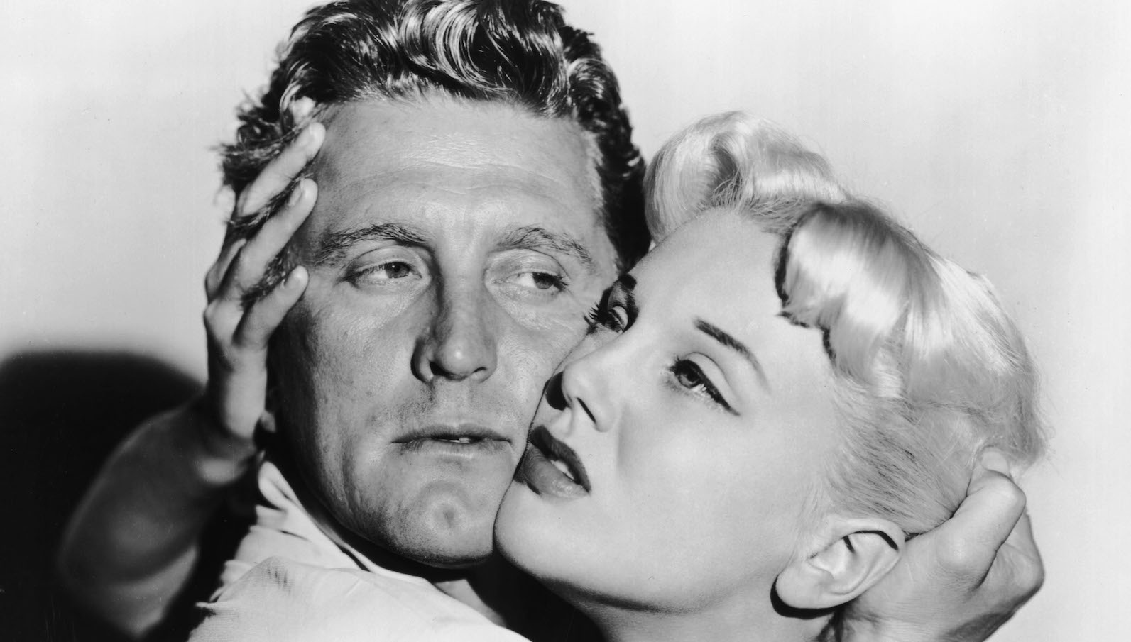 A man and woman hold each other's heads uncomfortably tightly as they look off into the distance in a black and white image