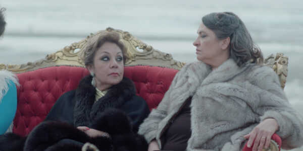 Two women in fur coats sit on a red sofa outdoors and look at each other