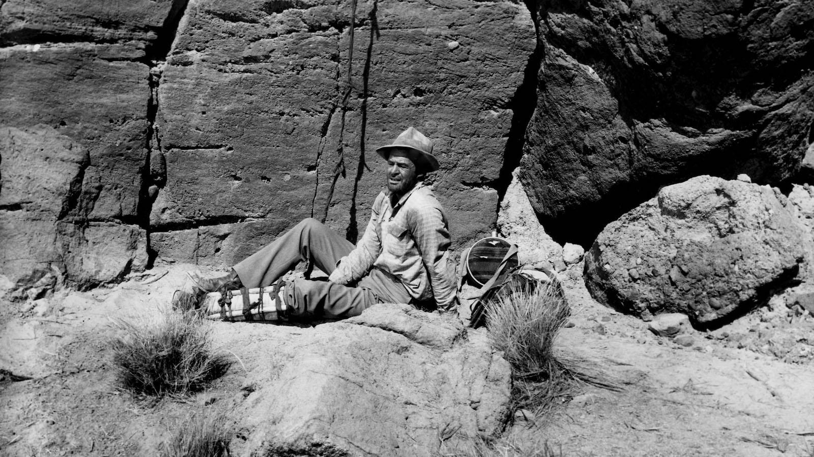 A man in a hat since in the desert by the side of a rocky mountain wall, holding his injured leg.