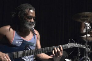 A man in a tank top with a white beard plays guitar