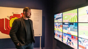 A man in a sports jacket stands admiring a grid of televisions showing a baseball game as it's edited live