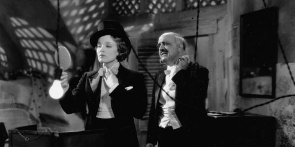 a woman in a tuxedo backstage ties her necktie and looks in a hand mirror, next to a mustachioed man