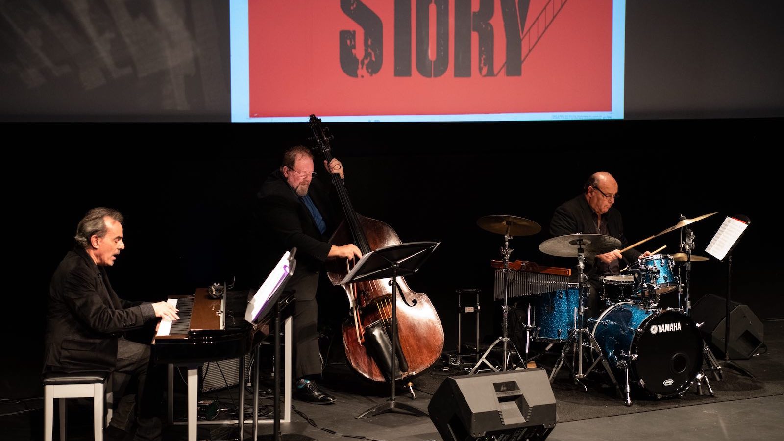 Three men play a piano, cello, and drums on a stage in front of a screen
