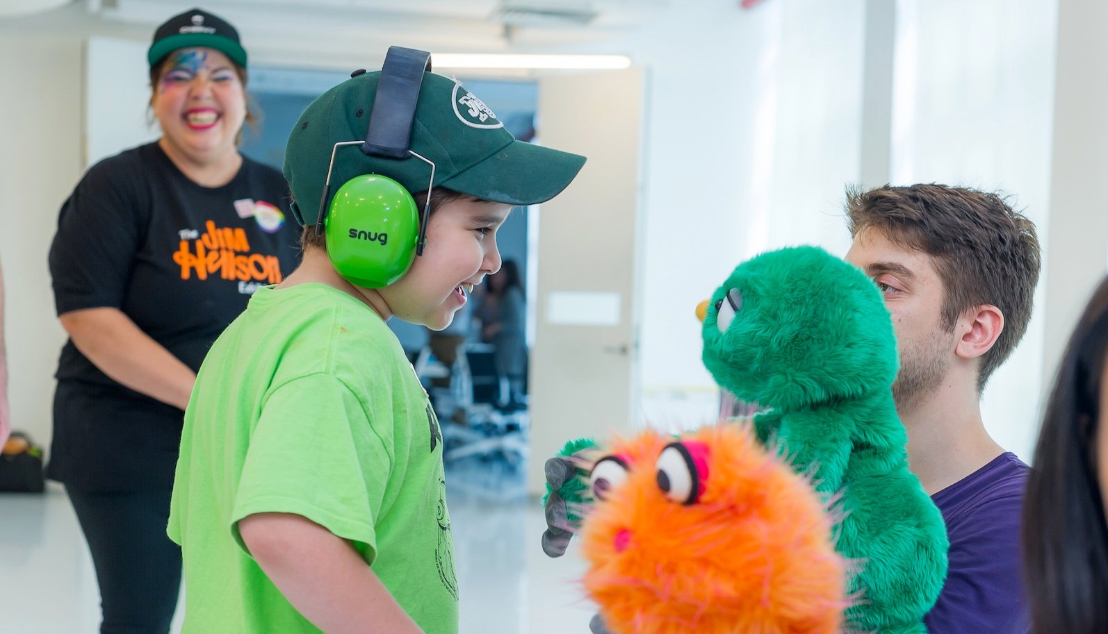 A little boy with green headphones and a green shirt interacts with a green puppet ...another orange puppet is in foreground.