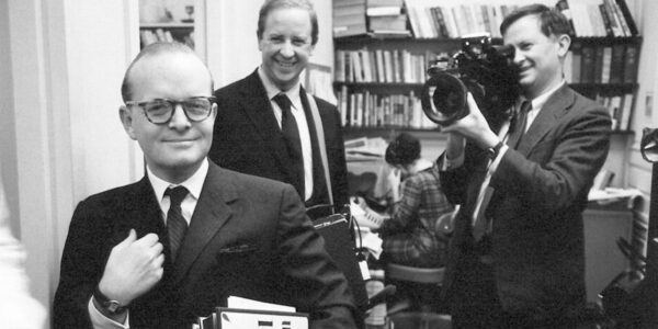 A man in a sharp suit and spectacles holds his lapel as two men behind him film him with a camera