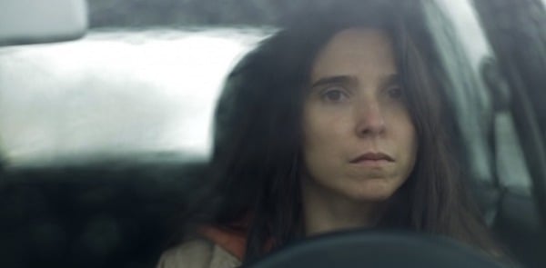 A dark-haired woman with a sullen expression sits at the wheel of her car behind smudged windshield glass.