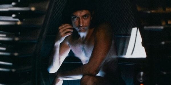 A naked man crouches in a futuristic machine pod, intensely looking at camera with one hand near his cheek